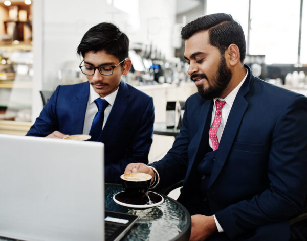 Two indian business man in suits sitting at office on cafe, looking at laptop and drinking coffee.