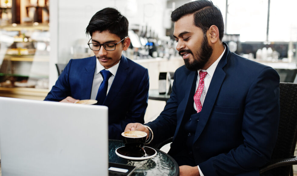 Two indian business man in suits sitting at office on cafe, looking at laptop and drinking coffee.